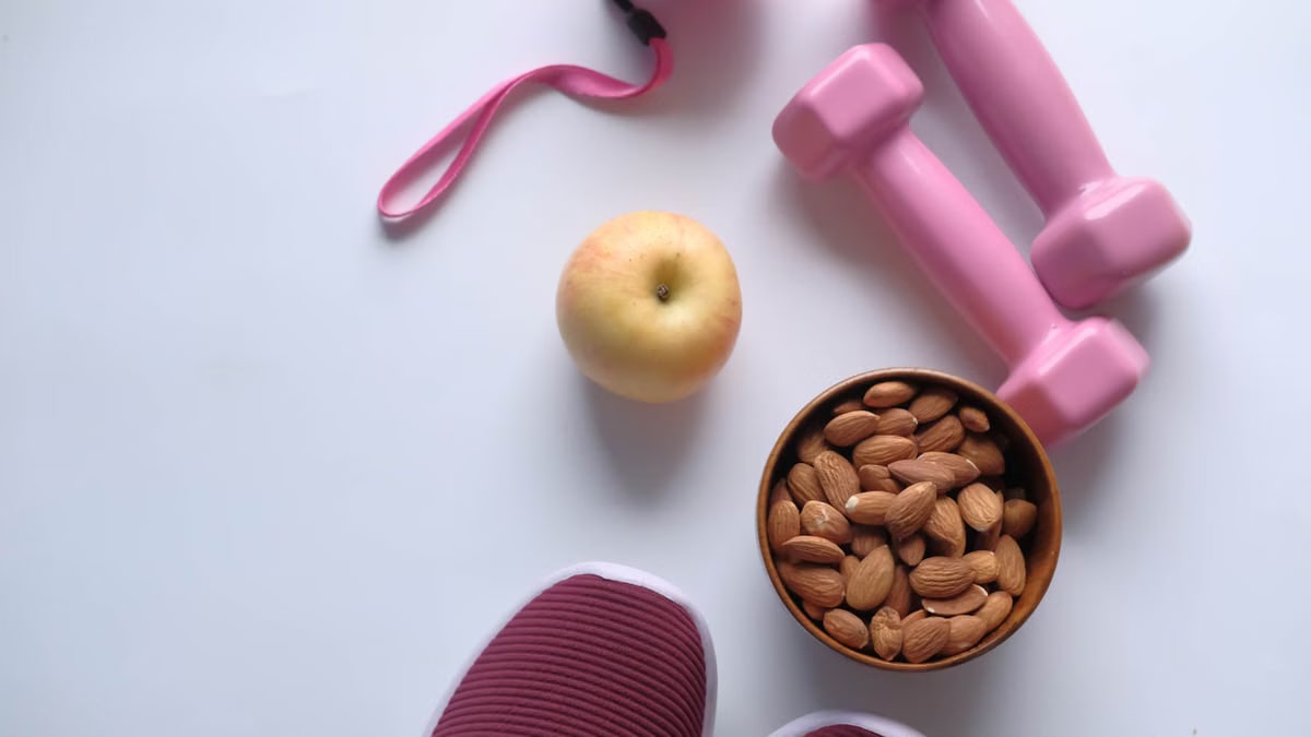 Flat lay of fitness essentials: pink dumbbells, almonds, apple, and sneakers on a white background.